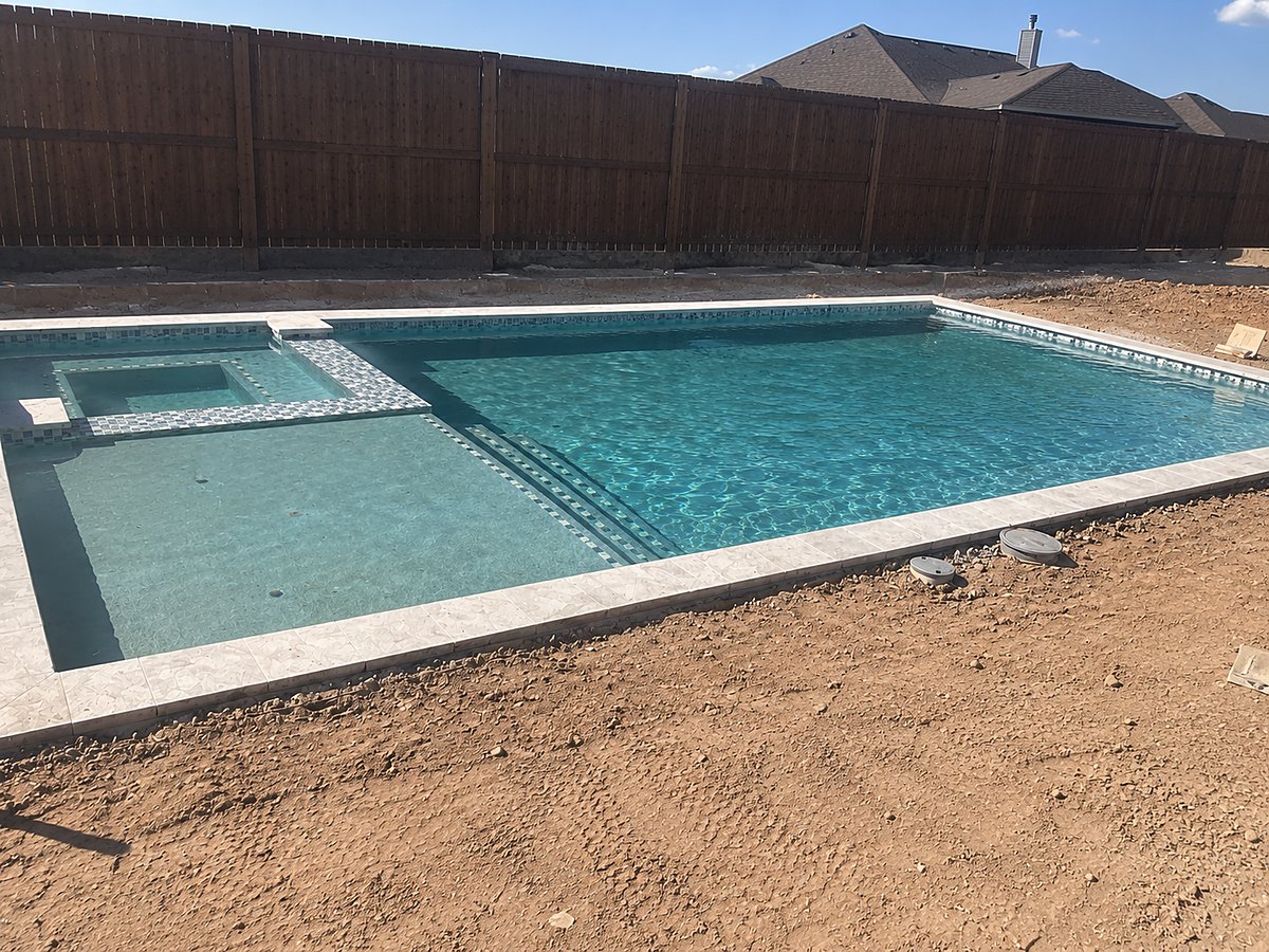 Pool and spa under construction with tile and travertine coping installed, behind a stained wood fence